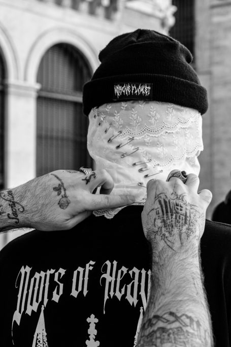 Hombre con tatuajes y una bufanda blanca cubriendo su rostro, vistiendo camiseta negra.