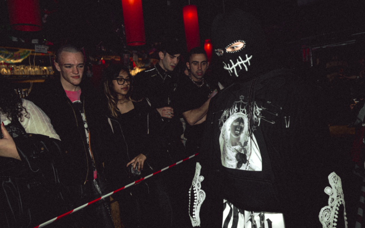 Persona con disfraz oscuro y máscara, rodeada de un grupo de espectadores.