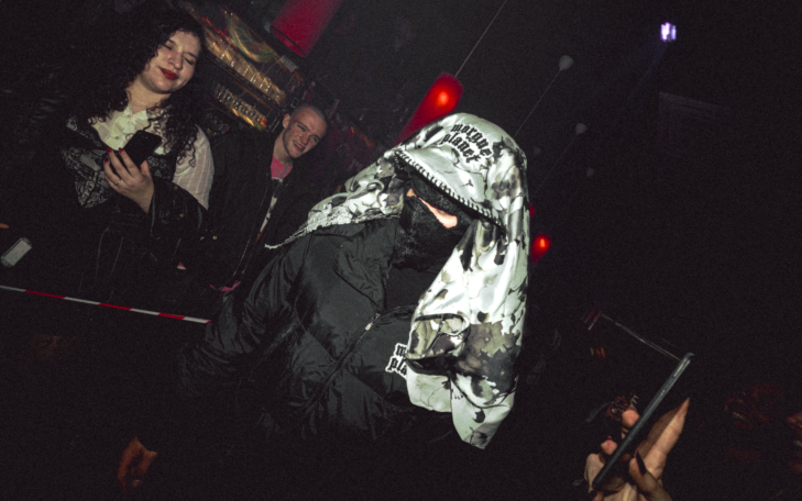 Persona con abrigo negro y un cubrecabezas blanco, rodeada de otras personas en un evento.