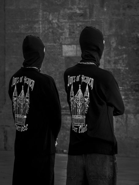 Dos personas de espaldas, vestidas con sudaderas negras y pasamontañas, en un entorno urbano.