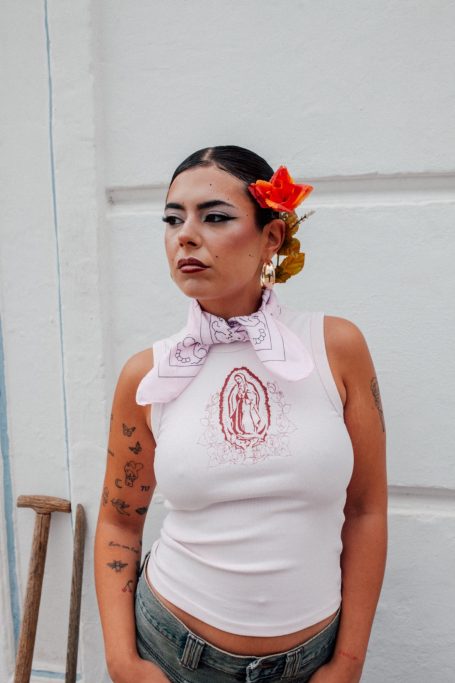 Mujer con top rosa, pañuelo en el cuello y flores en el cabello, posando ante una pared blanca.