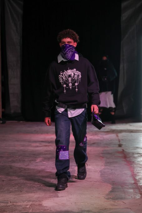 Modelo masculino con sudadera negra y máscara, caminando por una pasarela.
