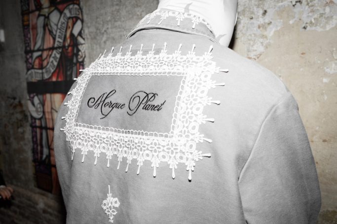 Modelo de espaldas con chaqueta gris de trabajo de Morgue Planet con lettering bordado en el centro y encuadrado con puntilla cosida.