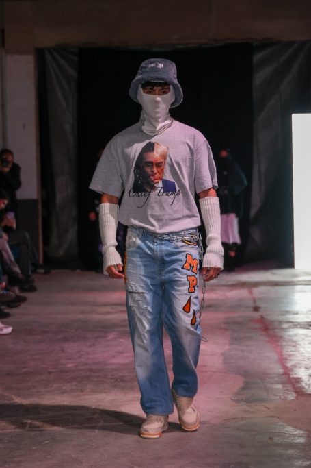 Modelo en pasarela con camiseta gris, pantalones vaqueros deshilachados y sombrero.