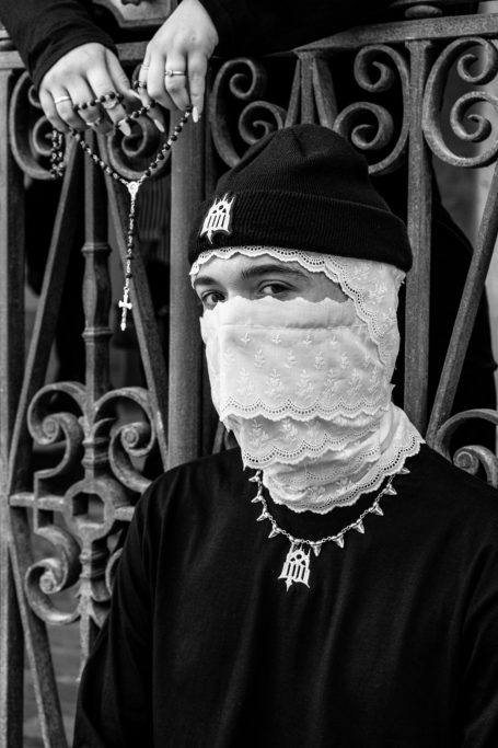 Joven con la cara cubierta y un gorro, sentado frente a una barandilla decorativa.
