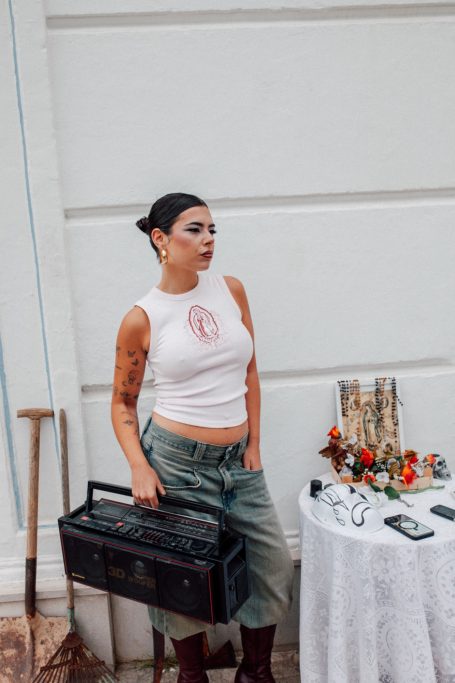 Joven con camiseta blanca y pantalones anchos, sosteniendo una caja negra junto a una mesa decorada.