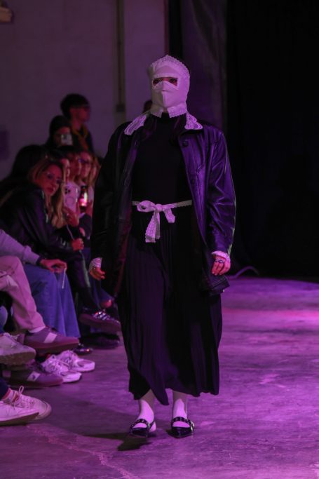 Modelo en pasarela, vestido con abrigo negro y máscara, presenta una colección de moda.