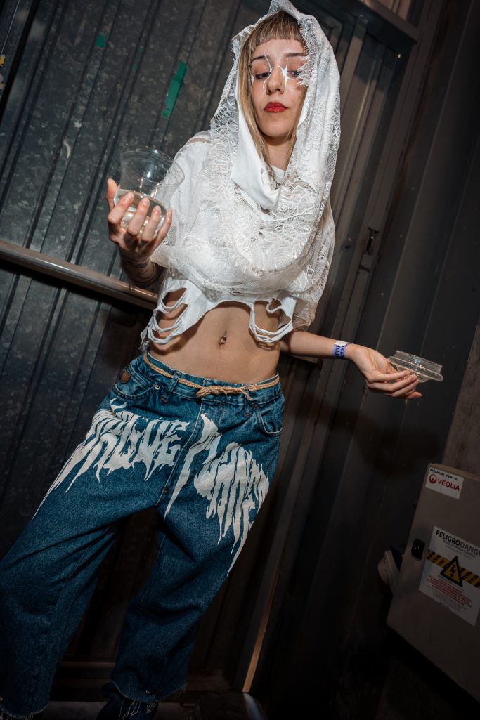 Joven con un atuendo moderno, blusa blanca y pantalones azules, posando de manera artística.