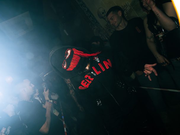 Persona con máscara y camiseta roja en un ambiente oscuro, rodeada de fans.