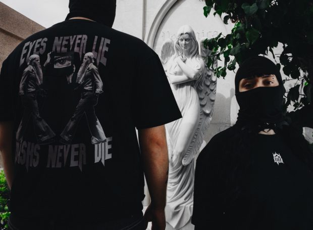 Dos personas con mascarillas negras frente a una estatua de ángel, una lleva una camiseta con texto.