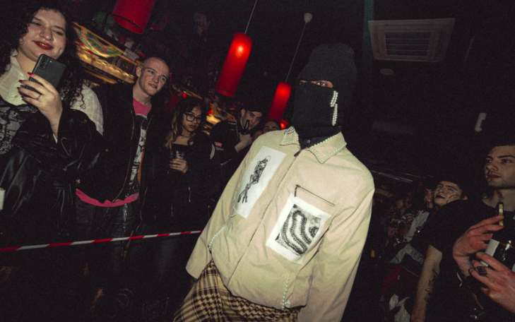 Modelo con máscara negra y ropa beige en un evento nocturno con público alrededor.