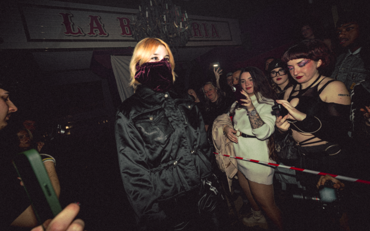 Persona con cabello rubio y atuendo oscuro en un ambiente de fiesta, rodeada de otras personas.