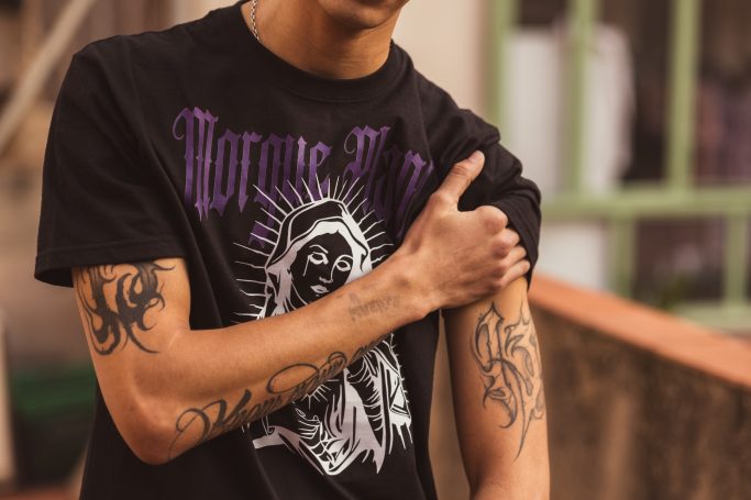 Joven con camisa negra y tatuajes en los brazos, mostrando uno en el brazo izquierdo.