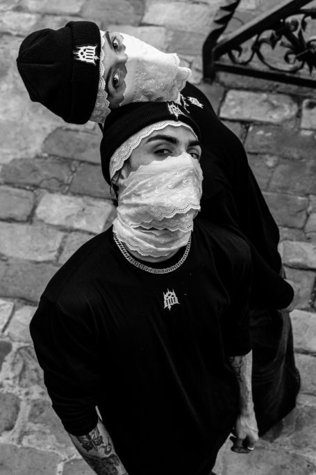 Dos personas con gorros y mascarillas, posando con una mirada seria en un entorno urbano.